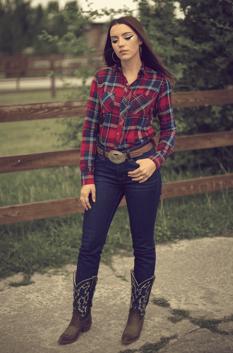 Stylish young woman in contemporary Western wear poses by a rustic fence outdoors.