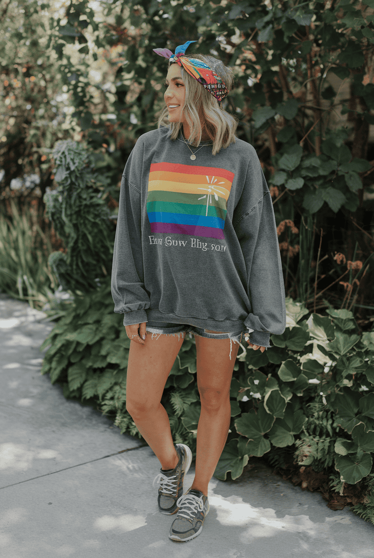 Woman in oversized rainbow sweatshirt and denim shorts smiles in a sunny outdoor setting.