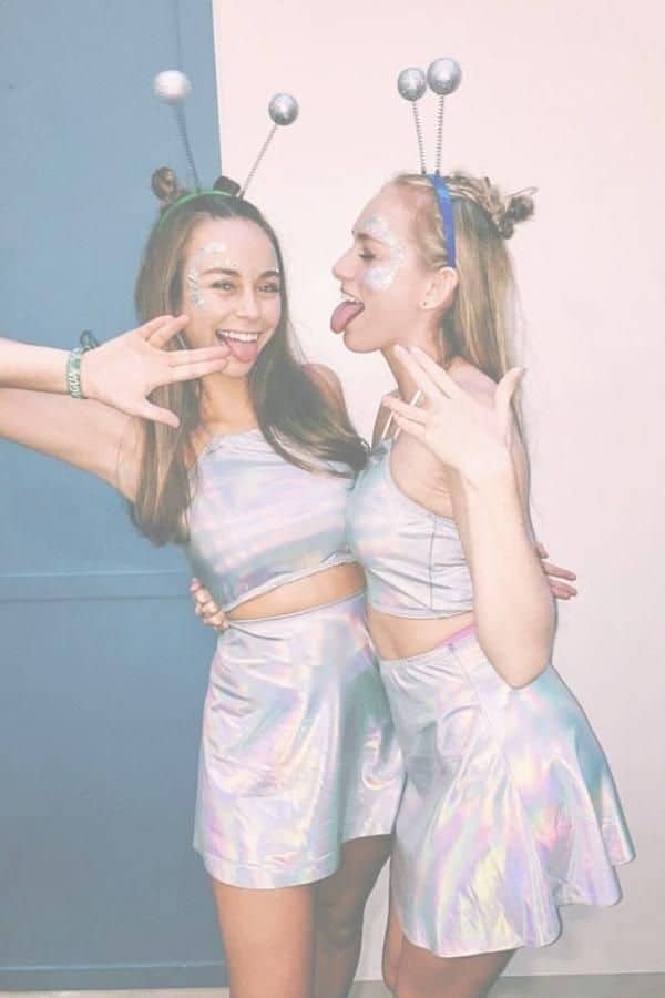 Two women in iridescent alien costumes with antenna headbands at a futuristic themed party.