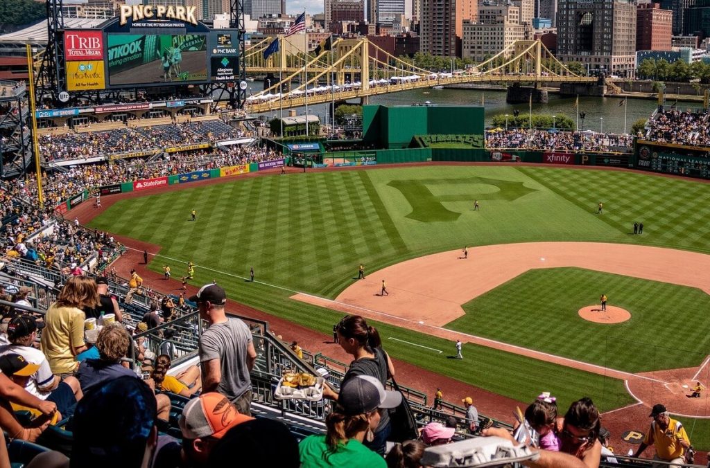 Fans in Pirates gear enjoy a sunny baseball game at PNC Park with the Pittsburgh skyline.
