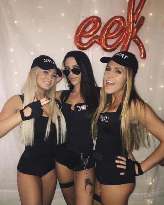Three college friends in matching SWAT Halloween costumes pose at a festive party with decorations.