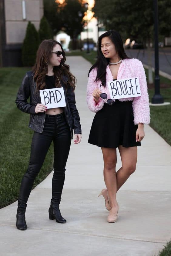 Two women in Bad and Boujee Halloween costumes pose confidently outdoors on a sidewalk.