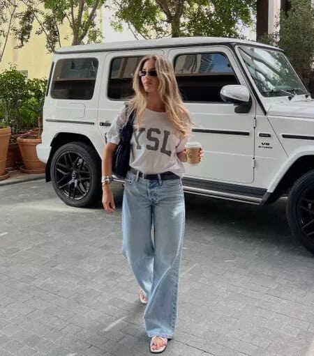 Trendy woman in YSL shirt and jeans walking by luxury white SUV, stylish street fashion.