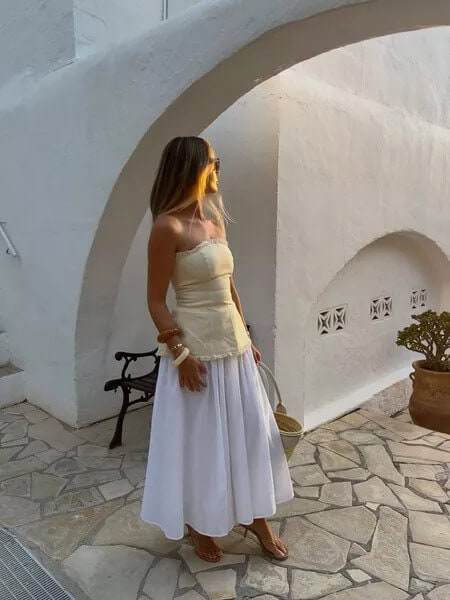 Woman in chic summer outfit with white skirt, Mediterranean architecture, sunset, stylish accessories.