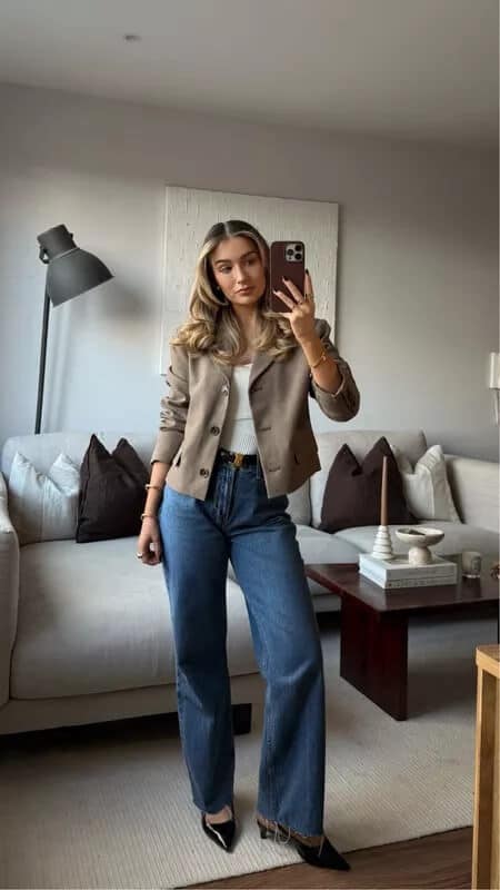 Woman in chic beige blazer and jeans taking a selfie in stylish modern living room.