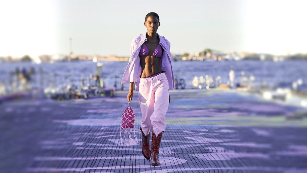 Model in purple bikini top, pink pants, red cowboy boots walking on colorful runway over water.