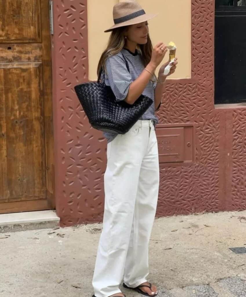 Stylish woman in striped shirt and wide-leg white pants enjoying ice cream on a sunny urban street.