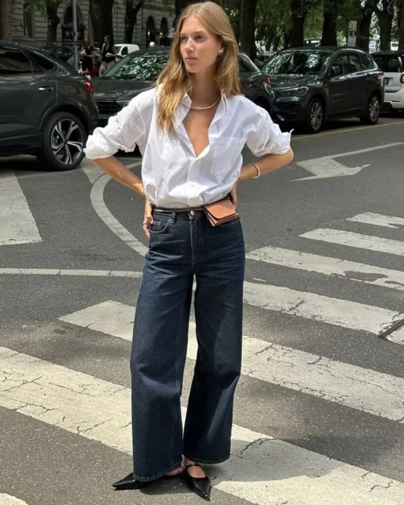 Trendy woman in oversized white shirt and wide-leg jeans crossing city street, summer street style.