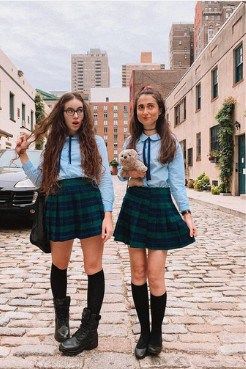 Two young women in plaid skirts and knee-high socks pose on a cobblestone city street.