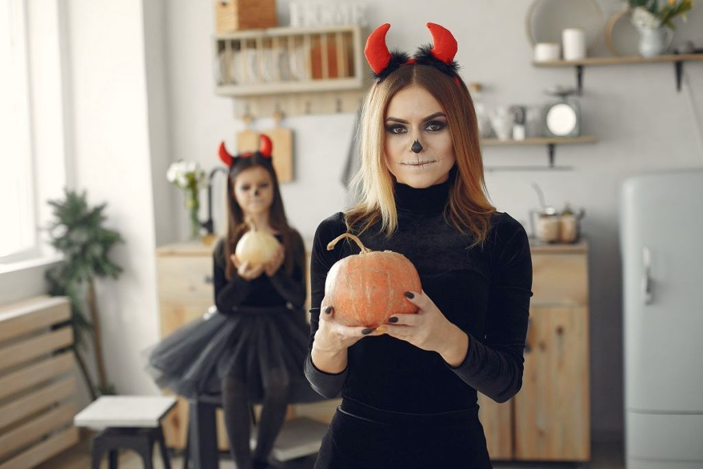 Women and girls in Halloween costumes with skull makeup holding pumpkins in a stylish home.