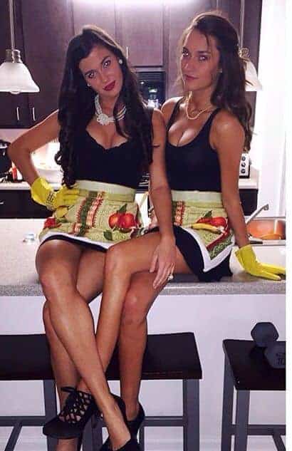 Trendy best friends in matching playful kitchen aprons, chic black outfits, and pearl necklaces.