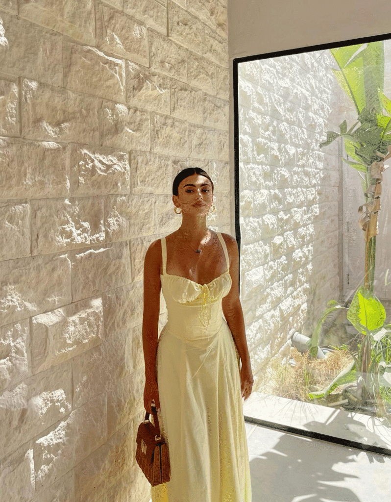 Woman in yellow summer dress with brown handbag stands by rustic stone wall in sunlit room.
