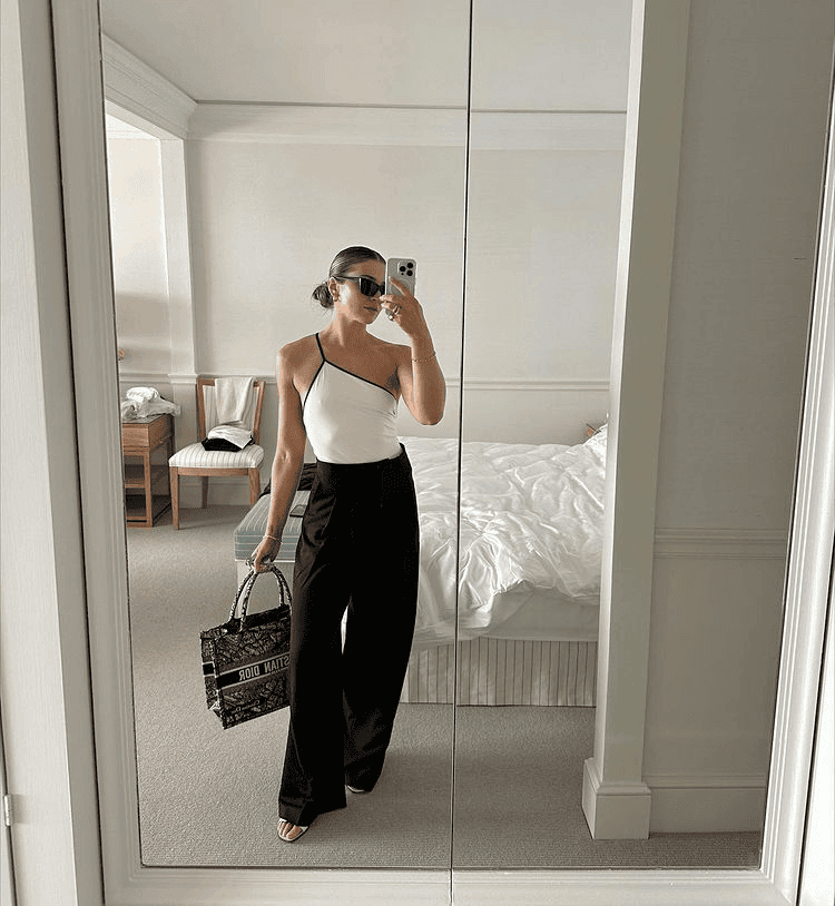 Chic woman in trendy black and white outfit posing in mirror with stylish bag and sunglasses.