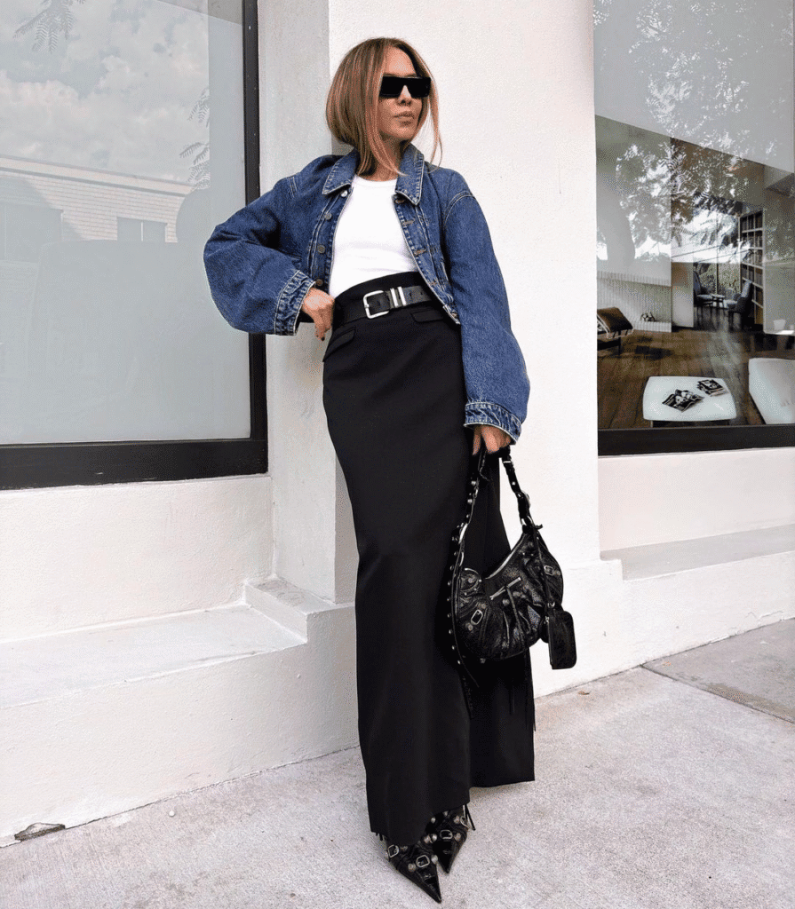 Chic woman in denim jacket, black maxi skirt, and sunglasses poses in modern urban setting.