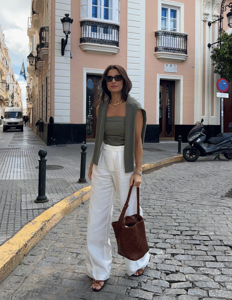 Chic woman in white trousers and cardigan on stylish European cobblestone street with pastel buildings.