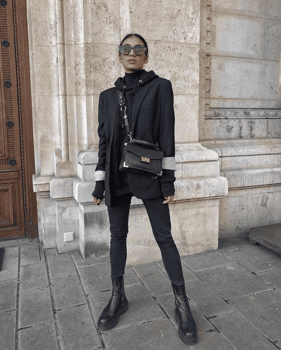 Stylish woman in black blazer and boots posing by historic stone wall, modern urban fashion.