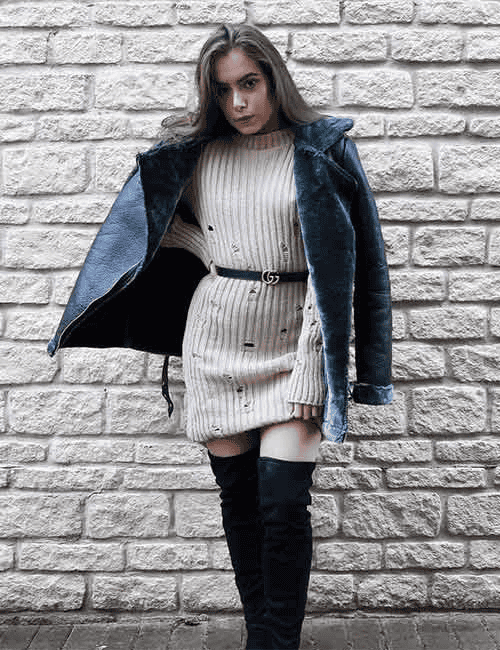 Stylish woman in knit dress and leather jacket posing by rustic stone wall in fall fashion.
