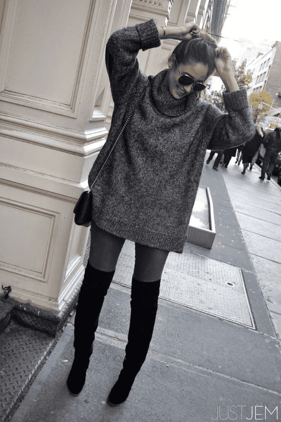 Stylish woman in oversized gray sweater, thigh-high boots, and sunglasses on a city street.