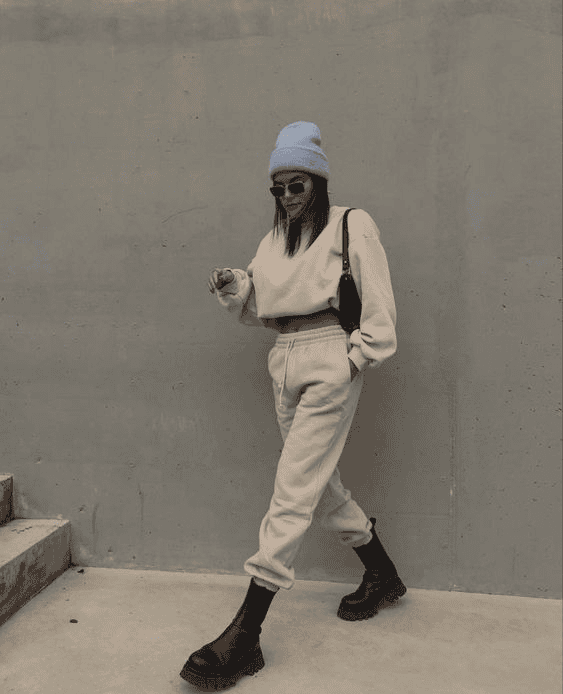 Trendy woman in beige loungewear, blue beanie, and combat boots walking by concrete wall.