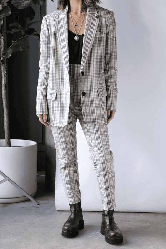 Woman in plaid suit and black boots poses stylishly in modern office with plant decor.
