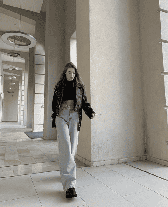 Stylish woman in modern architecture wearing black leather jacket, cropped sweater, high-waisted jeans, platform boots.
