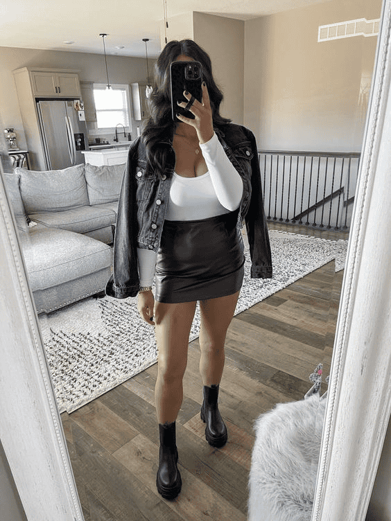 Woman in white top, brown mini skirt, denim jacket, black boots takes mirror selfie in modern living room.