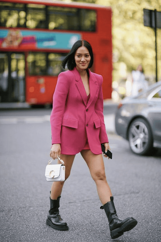 Stylish woman in pink blazer and shorts walks city street with combat boots and handbag.