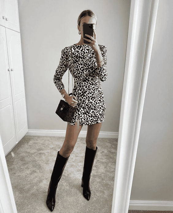 Woman in leopard print dress and black over-the-knee boots poses in modern bedroom mirror.