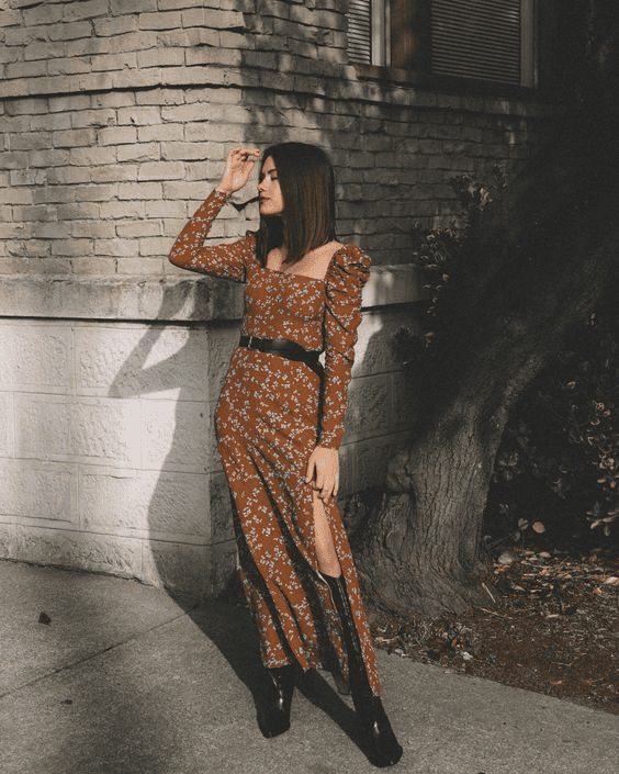 Woman in brown floral dress with black boots posing against a textured gray brick wall outdoors.
