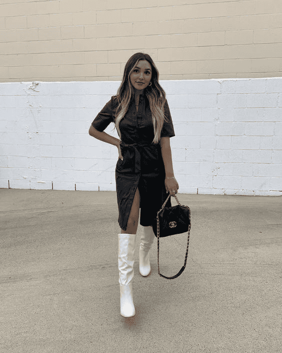 Stylish woman in black dress and white boots walking by white brick wall, modern urban fashion.