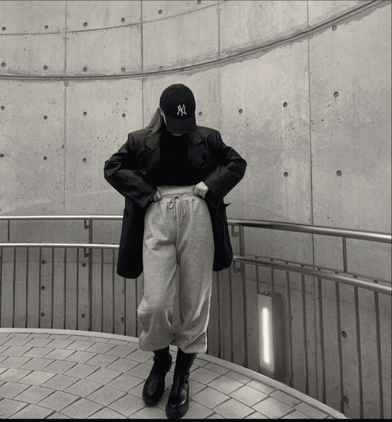 Stylish woman in modern minimalist concrete setting, wearing gray sweatpants, black coat, and cap.