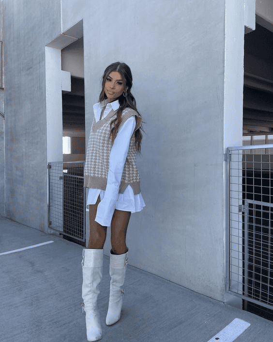 Stylish woman in houndstooth vest and white boots poses in modern urban setting.
