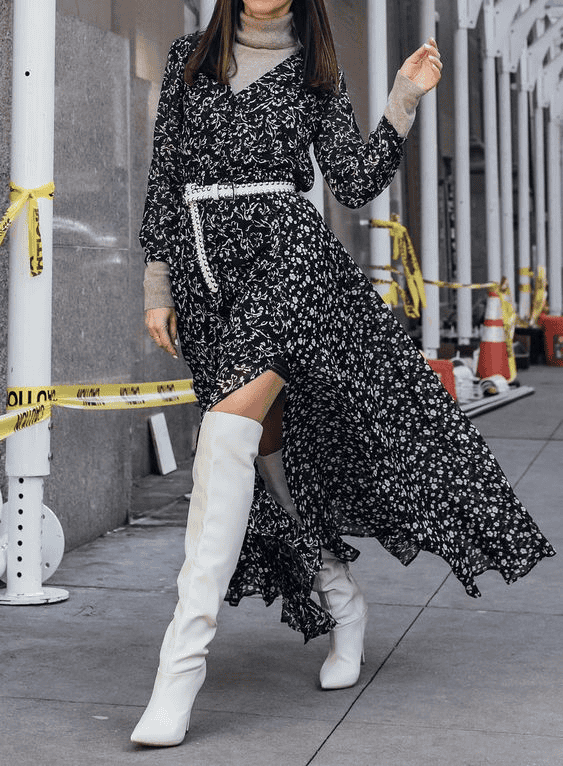 Fashionable woman in floral black dress and white boots poses in urban street with construction tape.