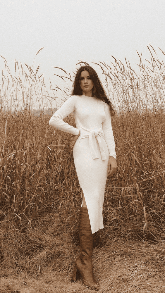 Woman in cream sweater dress and tall boots posing in autumn field under cloudy sky.