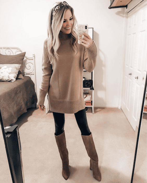Woman in beige sweater, black leggings, and tan boots posing stylishly in a cozy bedroom.