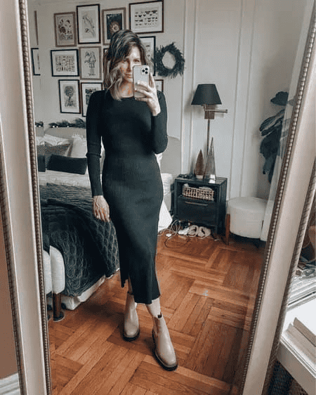 Woman in black ribbed dress taking mirror selfie in cozy, stylish, modern bedroom with decor.