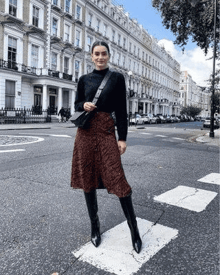 Stylish woman in black sweater and burgundy skirt crossing street with Victorian architecture background.