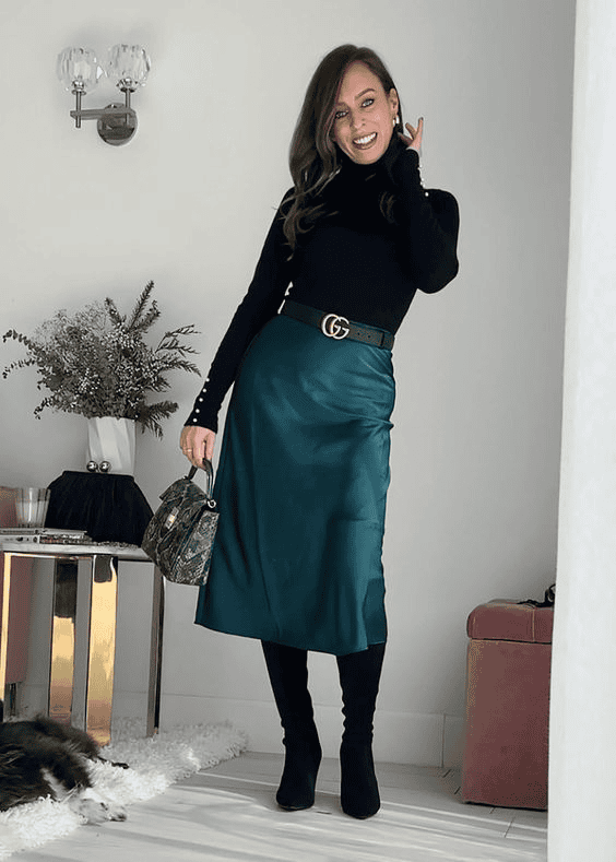 Stylish woman in teal satin skirt and black turtleneck poses in modern chic living room.