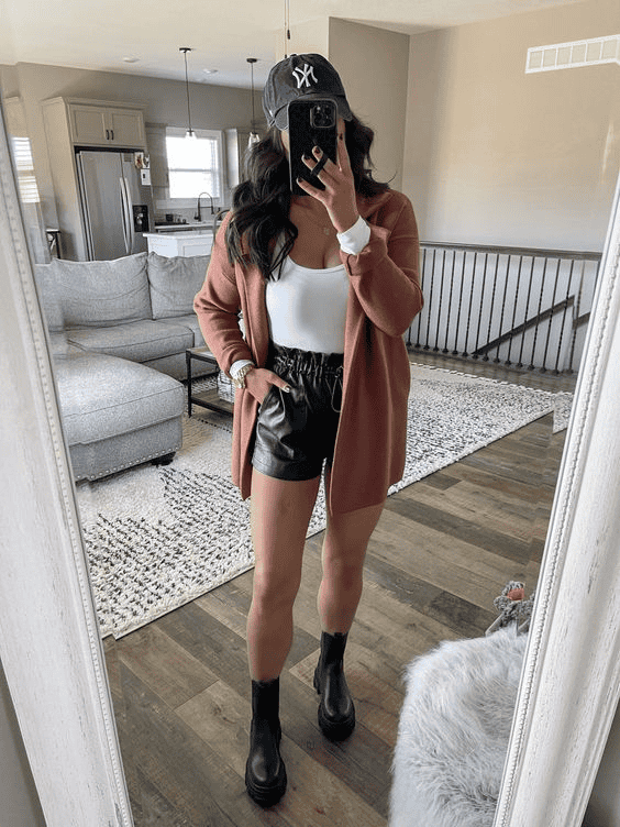 Trendy woman in faux leather shorts and pink cardigan taking a mirror selfie in modern living room.