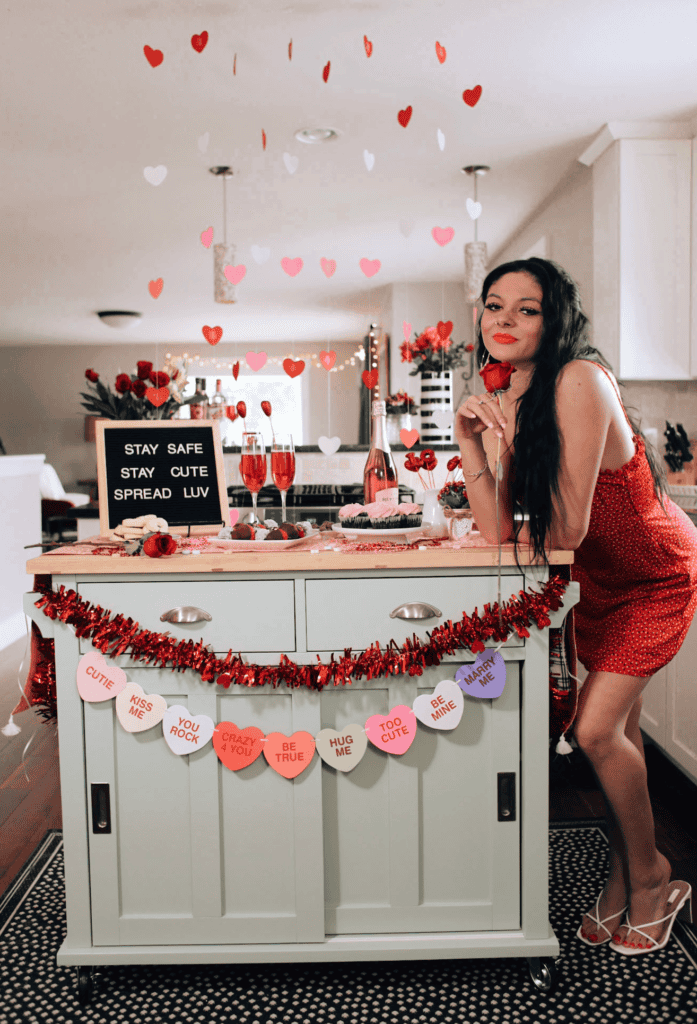 Woman in red dress celebrating Valentine’s Day in festive kitchen with hearts, champagne, and treats.