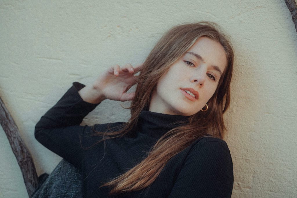Young woman in black turtleneck posing against a warm textured wall with natural lighting.