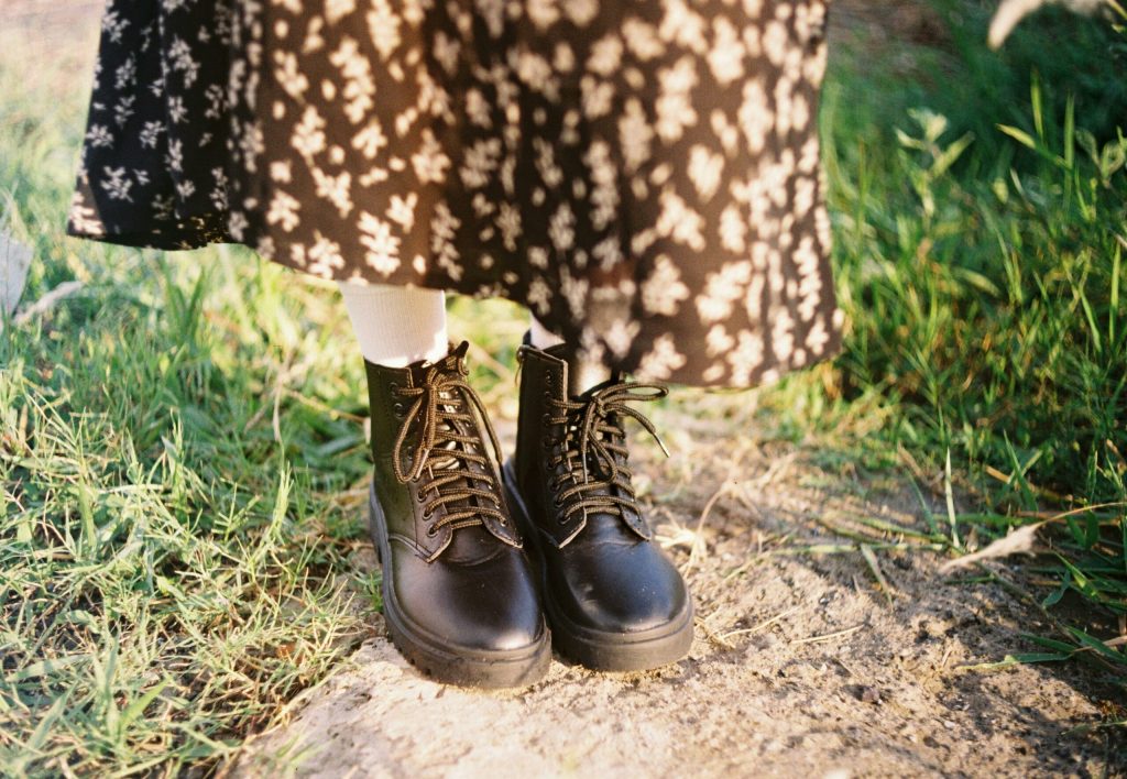 Woman wearing black boots, floral skirt, and white socks standing on grass outdoors in sunlight.