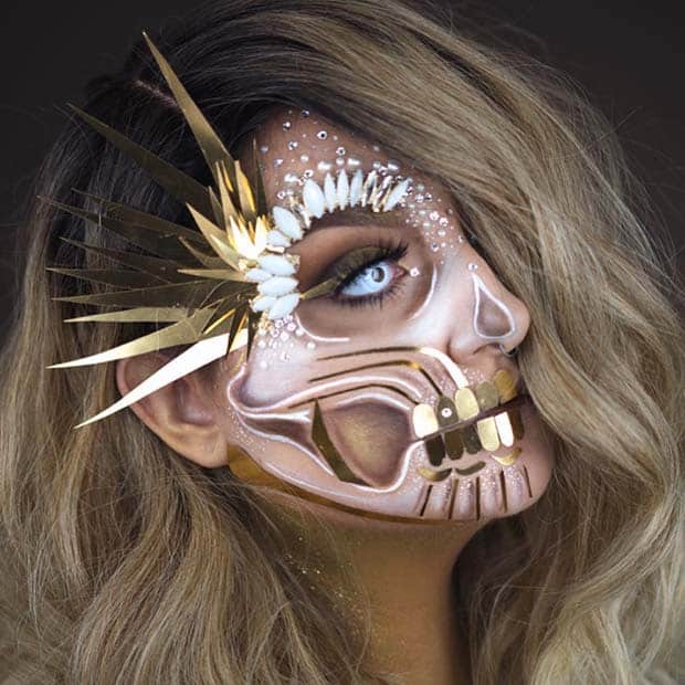 White and gold skull Halloween makeup on woman with blue eyes and metallic headpiece.