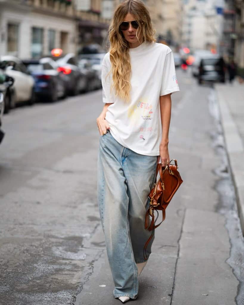 Trendy woman in oversized graphic tee and jeans walks city street, urban street fashion style.