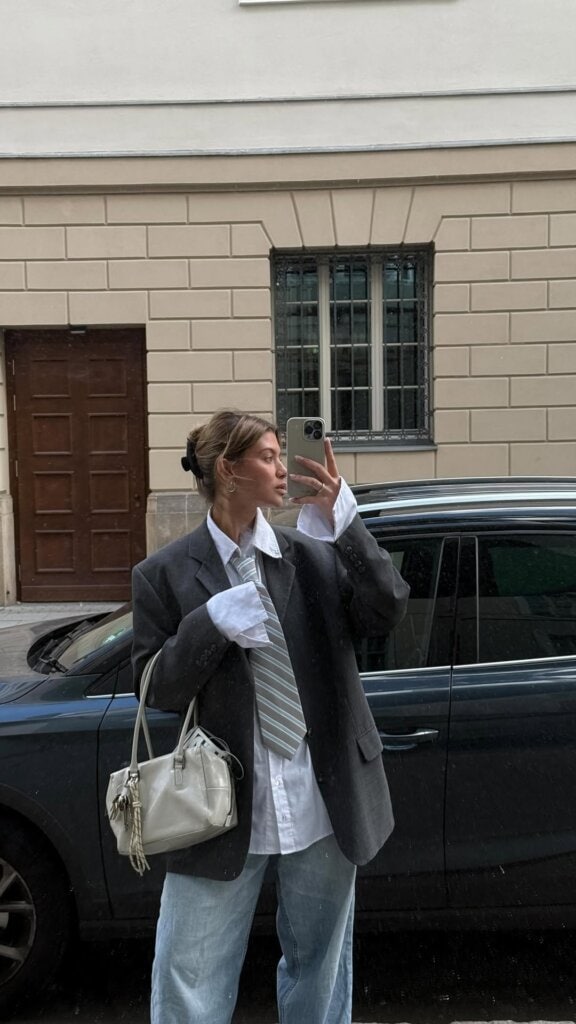 Young woman in oversized blazer and tie taking a street style mirror selfie in urban setting.