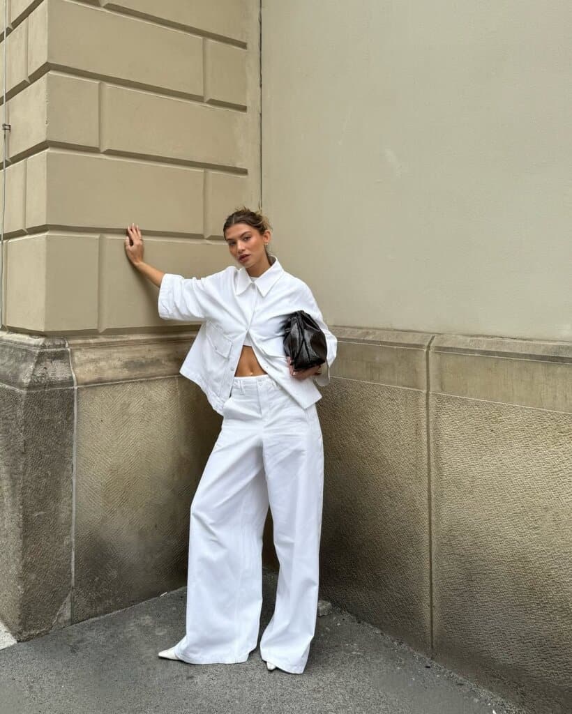 Chic street style: woman in all-white outfit with black clutch, urban fashion background.