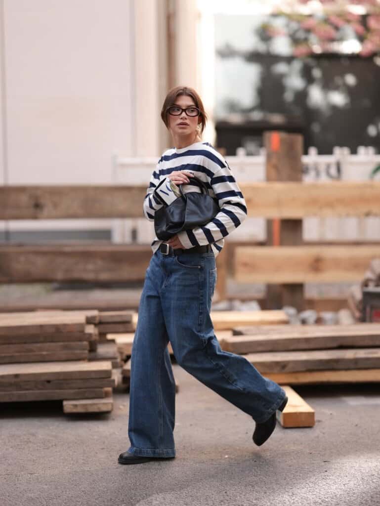 Trendy woman in striped sweater and jeans poses in urban street fashion setting.