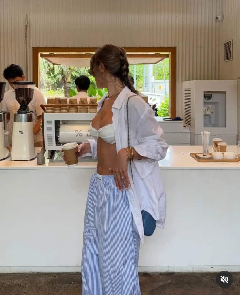 Trendy woman in streetwear, oversized shirt, bralette, striped pants at modern coffee shop.