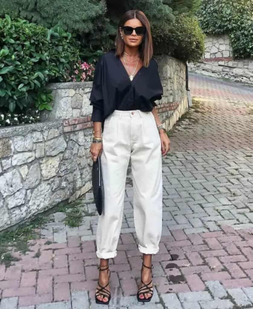 Chic woman in trendy streetwear with black blouse, cream pants, sunglasses, and strappy heels.