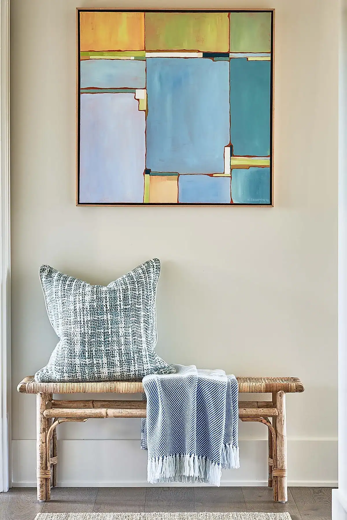 Rattan bench with a textured pillow and throw blanket beneath colorful abstract art in a modern entryway.